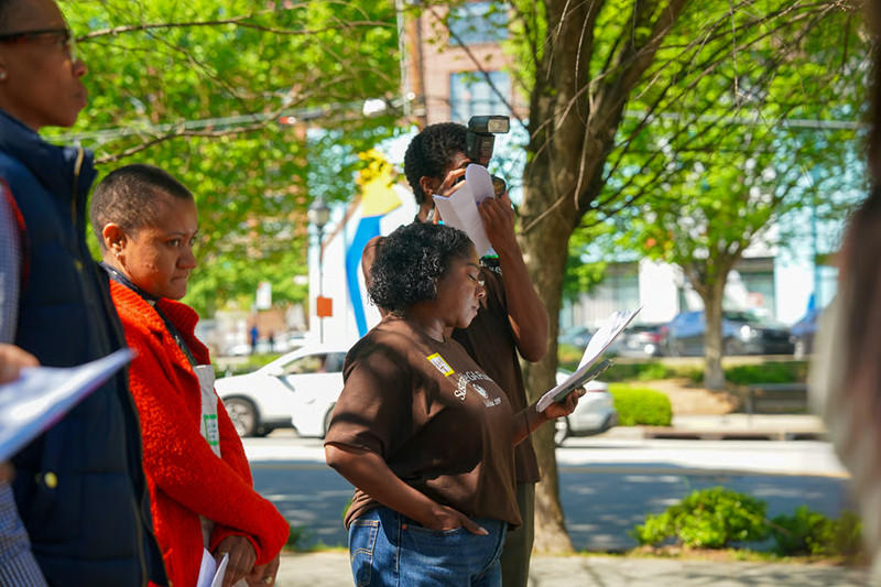 People standing on a tree-lined city sidewalk holding papers, with a photographer capturing the moment in the background.