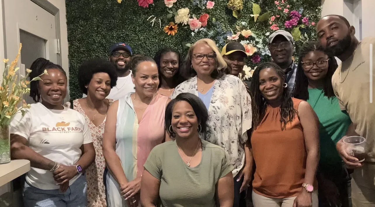 Group photo of about a dozen smiling people of varying ages standing and posed in front of a decorative flower wall backdrop