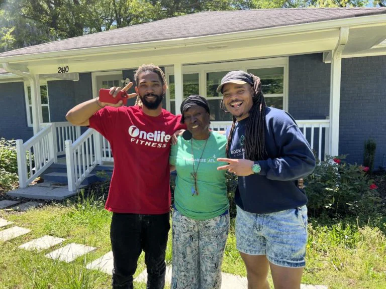 2 men standing on the outside of an older woman holding peace sign in front of a home